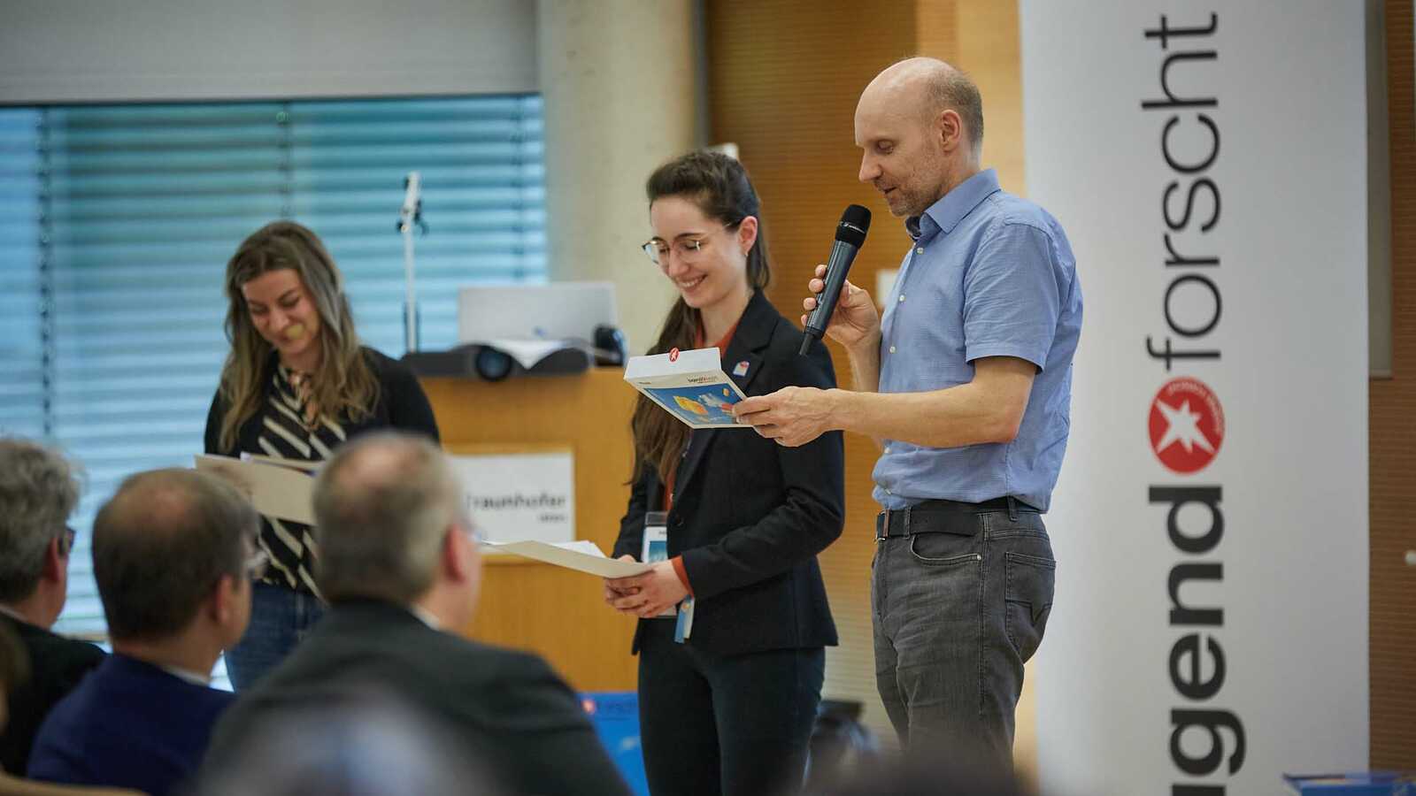 Jurymitglied hält Laudatio in Anwesenheit einer Preisträgerin und der Moderatorin